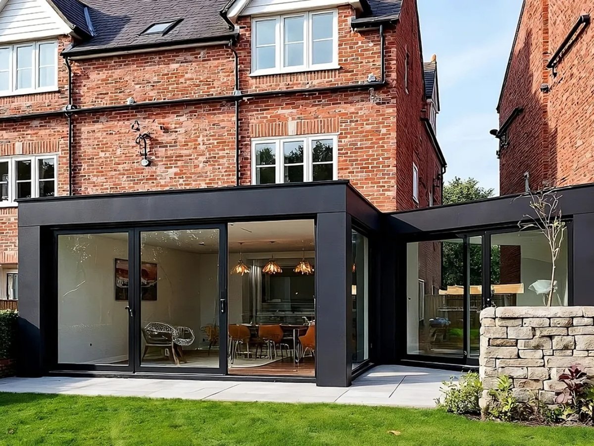 Modern side extension with large glass doors, showcasing a stylish dining area, set against a classic red brick house. Green lawn in foreground.