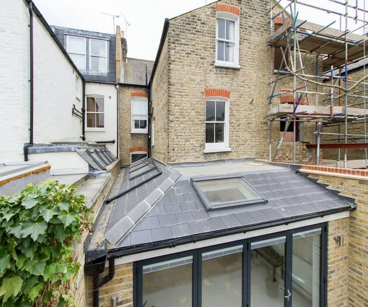 View of a small wraparound extension with a slate-roof and large windows, surrounded by brick buildings and construction scaffolding.