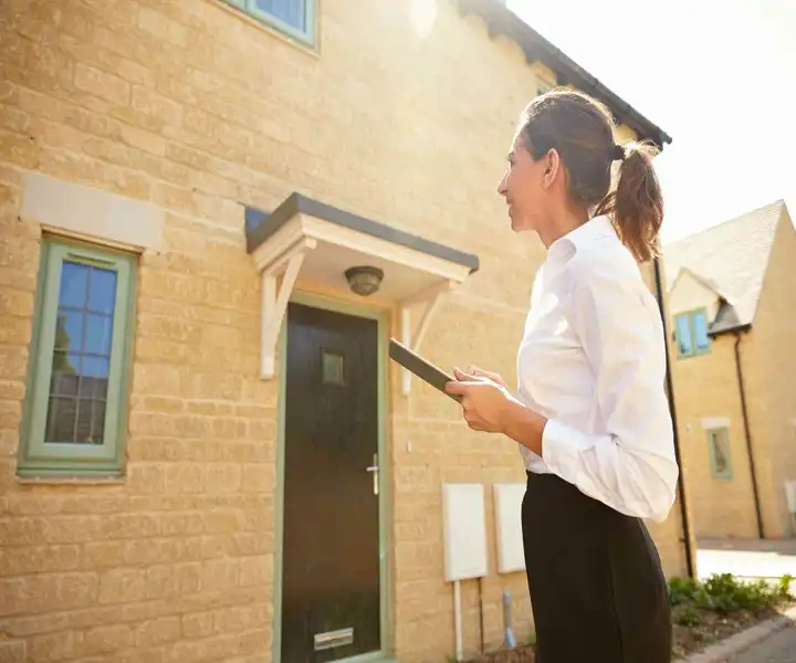 A woman in a white blouse stands in front of a modern stone house, holding a tablet and inspecting and surveying the entrance.